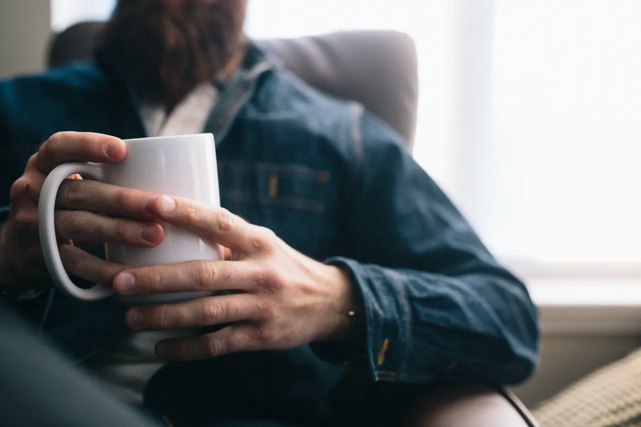 Bearded man in a denim jacket holding a white mug while seated in an armchair by a bright window